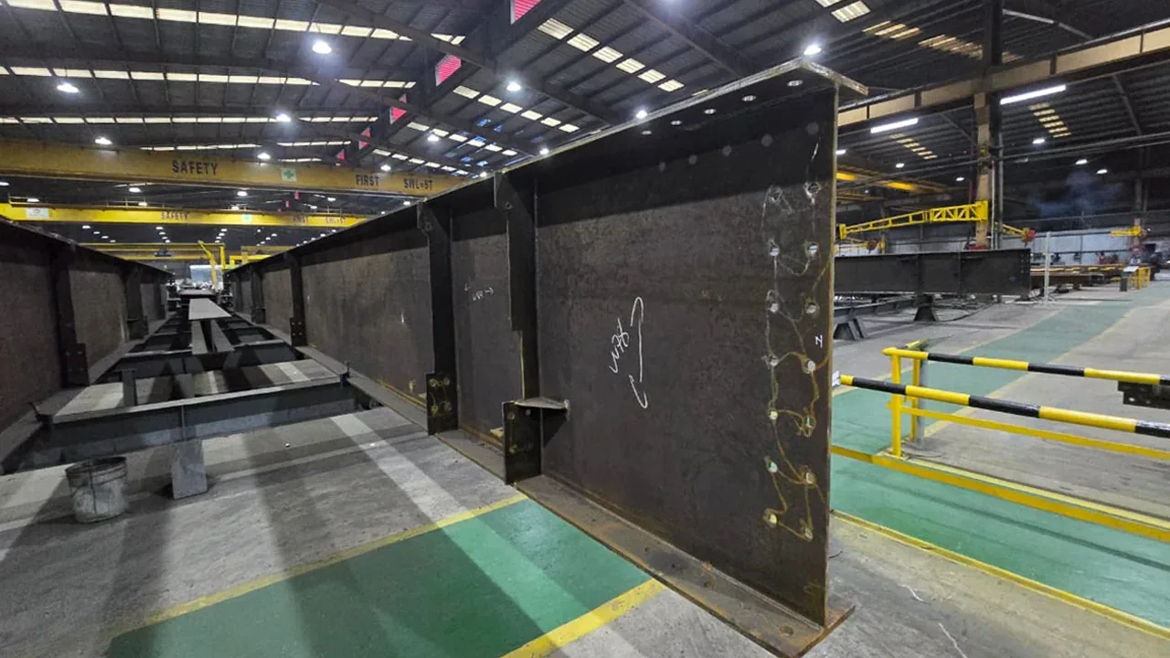 Built-up tapered steel girder on the fabrication shop floor with bolt holes drilled and layout marks chalked on the web