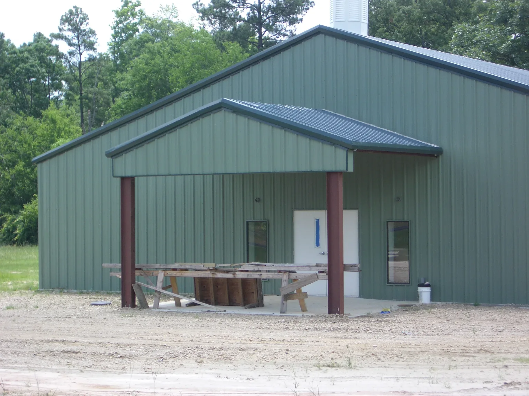 Green metal building with a gabled roof and a deep covered porch supported on steel columns