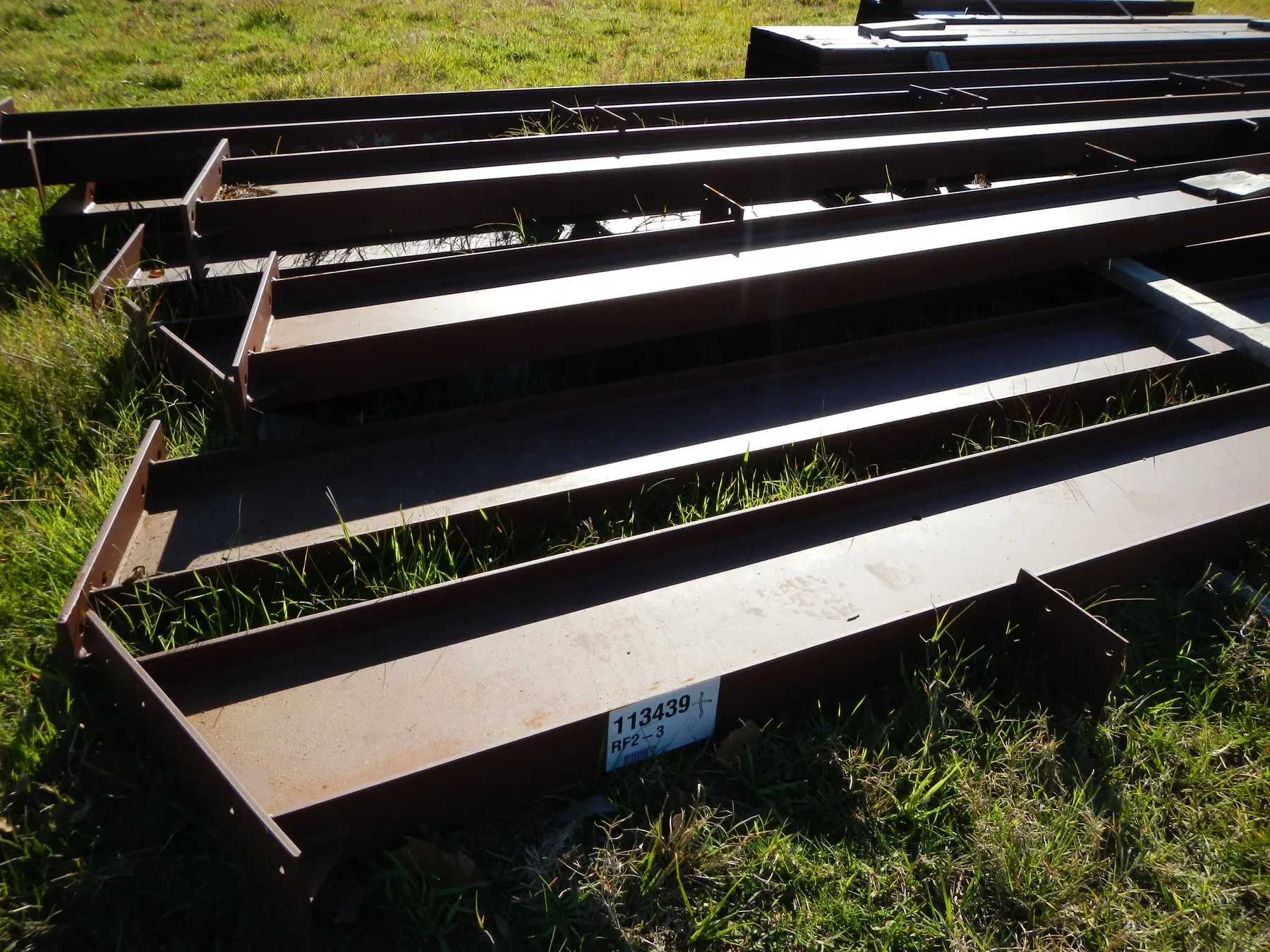 Stacked red-iron rigid frame members delivered to site and laid out on the grass before erection